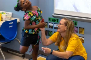 A young girl happily hands a picture to her teacher who is kneeling down and smiling