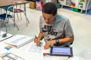 A young adult works on school work with his AAC device next to him